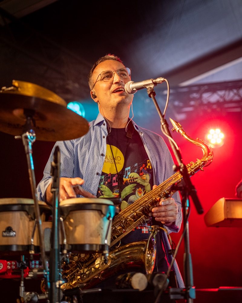 Bandmedlem synger og spiller percussion og saxofon p&aring; scenen