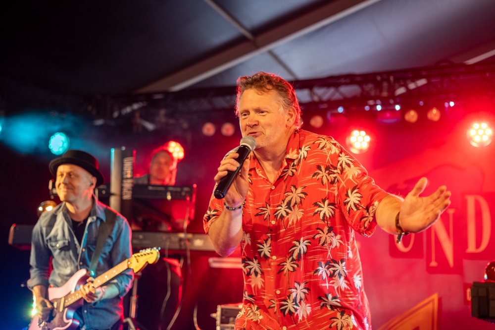Forsanger i r&oslash;d skjorte og guitarist p&aring; scenen med bandets banner i baggrunden